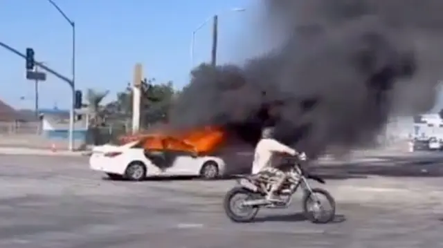 A car is seen on fire and a person on a motorcycle drives in front of it. It is broad daylight