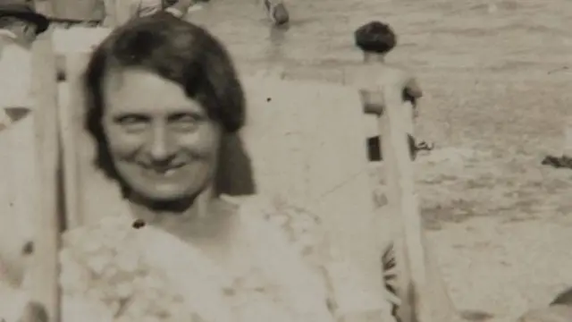 An old black and white photo of a woman. She is pictured sitting on a deck chair, looking at the camera and smiling.