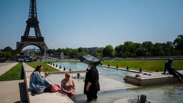 A woman stands in the water inside a fountain at the Trocadero in front of the Eiffell Tour, two women sitting down as one of them fans herself with an electrical fans