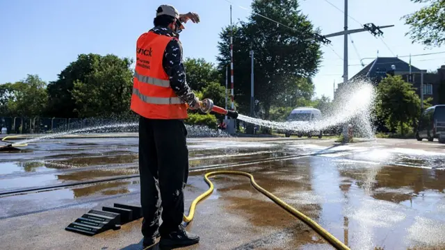 A city employee in orange hi-vi vest and black trousers sprays water on a bridge to cool the road surface during a heatwave in Amsterdam