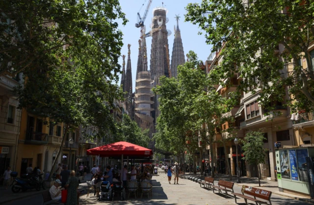 Barcelona's Sagrada Familia pictured from a tree-lined avenue with a row of wooden benches on the right side and tourists eating al fresco under the shade of a restaurant's red tent