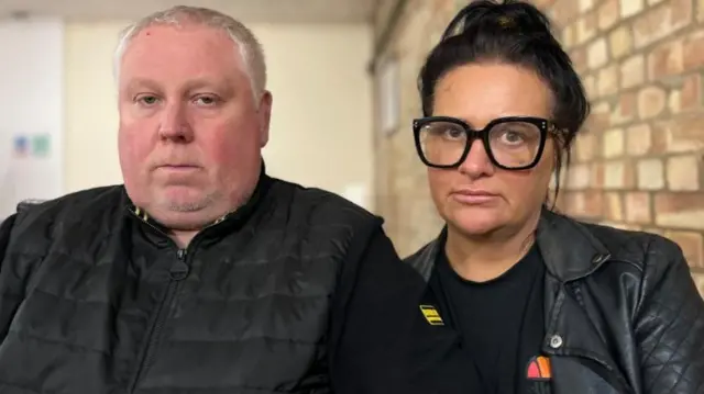 Man with white hair in a balck jacket sits close to a woman with long black haired tied up. She is wearing a leather jacket and large spectacles with a thick black frame. It is Mark and Caroline Willgoose speaking to the BBC after the death of their son.