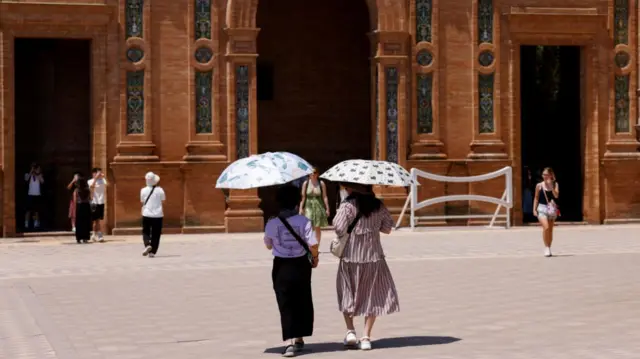 Two women wearing skirts and short-sleeved tops walk in Seville, other tourists milling around outside a red-stone building