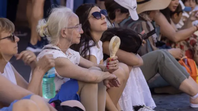 People sit on the side of a road and hold fans to try to cool themselves