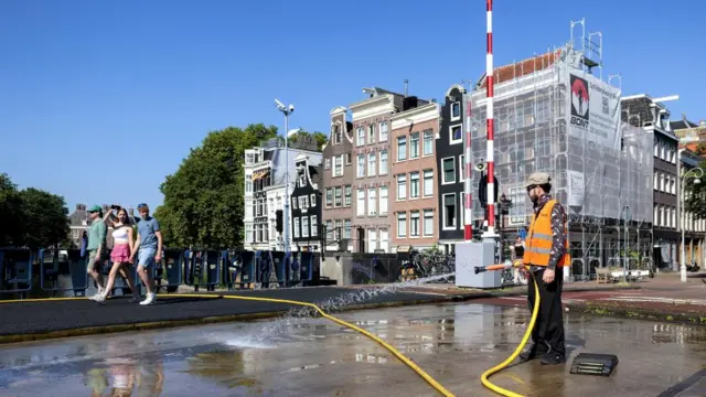 A man stands in the road, holding a hose spraying water onto the street. Three people walk past above canal, house line the right hand side.