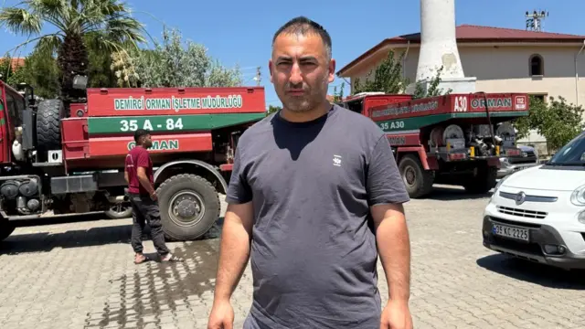 Galip Ener (man in short-sleeved grey shirt) stands in what appears to be a parking lot, a white car to his left and two red and green lorries behind him