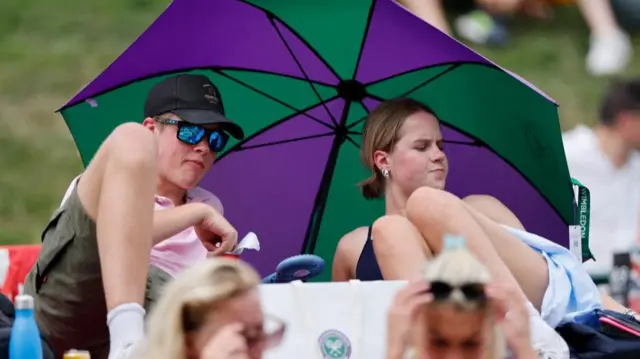 Tennis - Wimbledon - All England Lawn Tennis and Croquet Club, London, Britain - July 1, 2025 Spectators in the lawns take cover under an umbrella amid the heatwave