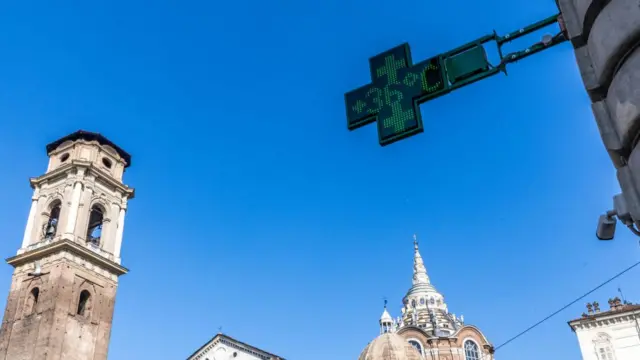 Top of a church bell tower to the far left of the frame, blue skies and the green cross-shaped temperature monitor of an Italian chemist's shop in Turin showing a 36C temperature