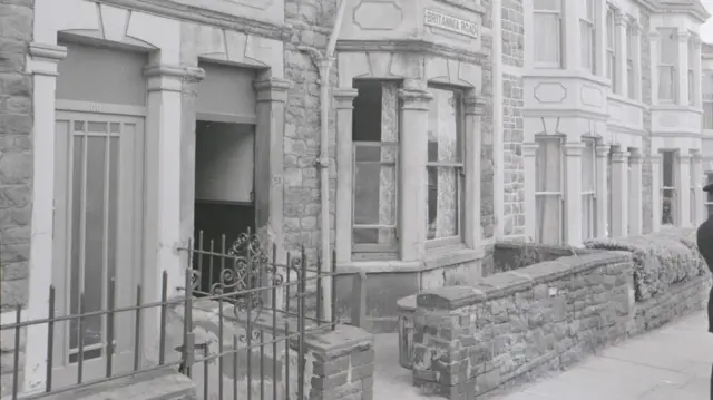 A black and white photo of a house. There is a small brick wall at the front of it.