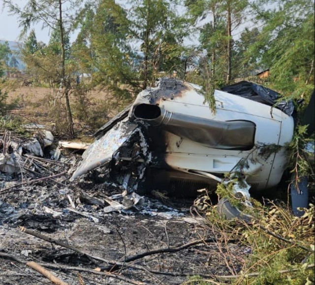 A plane is seen on the ground in a forest. The remains are charred out and it is split into pieces.