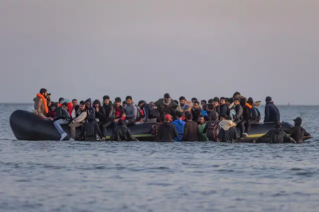 People thought to be migrants sit atop of a dingy in the open water. Some are wearing life vests. It is predominantly a group of about 30 young men.