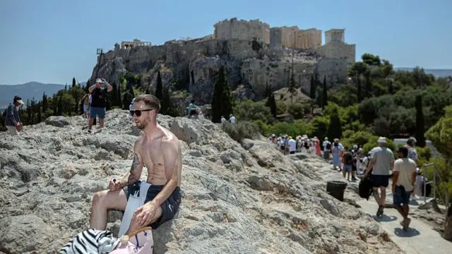 A shirtless man sits on rocks on Aeropagus hill in Athens, the ancient Acropolis visible on top on nearby hill surrounded by trees, tourists walking around