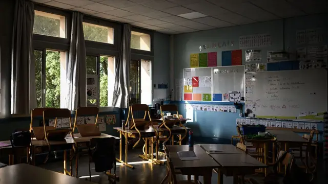 An empty classroom in a school in Bordeaux, France, with wooden and metal chairs piled up on wooden desks, lights are turned off. The wall is decorated with colourful informative paper and two boards