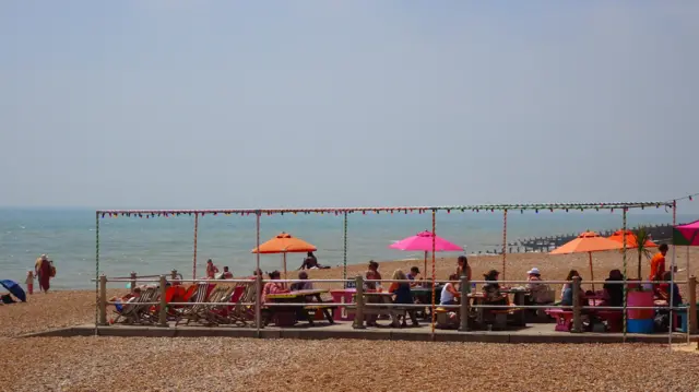 People on a platform on a beach sat at tables with sea behind and umbrellas up.