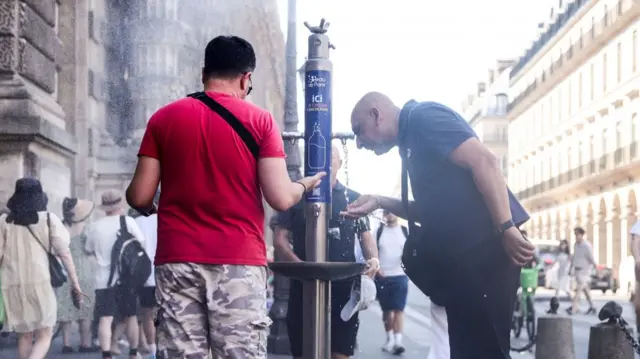 A man in a red top and camo trousers drinks from a Paris water fountain, a bald man in a dark polo shirt and long black trousers drinking next to him