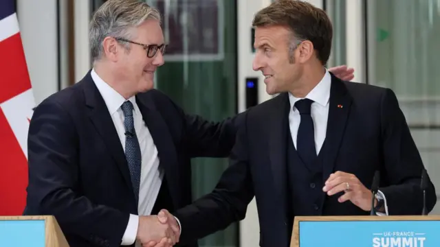 Starmer and Macron stood behind podiums side by side, they shake hands and look at each other. Both wearing dark suits.