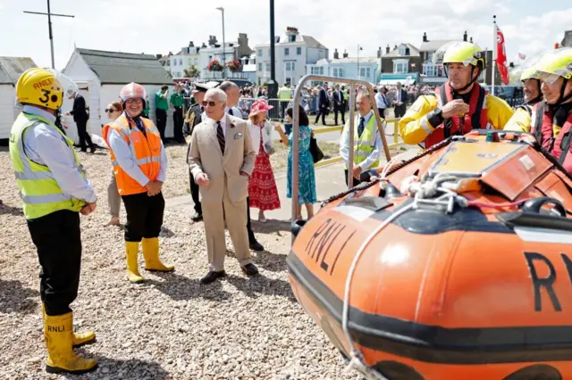 King Charles speaks to RNLI volunteers, two are wearing high vis vests, helmets and yellow wellington boots. Three are wearing their rescue gear, life vests, helmets and are next to an orange lifeboat.