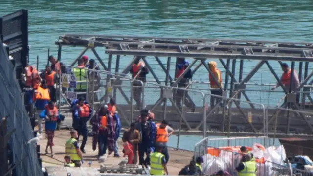A group of people, some in life jackets, walking along a metal gangway onto a port