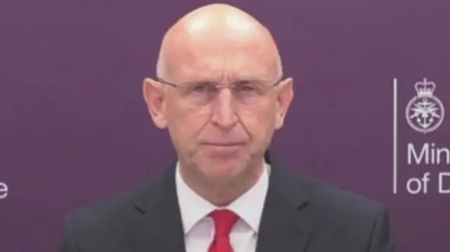 Headshot of Healey who wears a suit and shirt and red tie, as well as thin framed glasses. He sits in front of a purple background