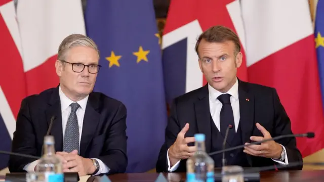 Starmer and Macron speaking at a table in front of EU, French and British flags