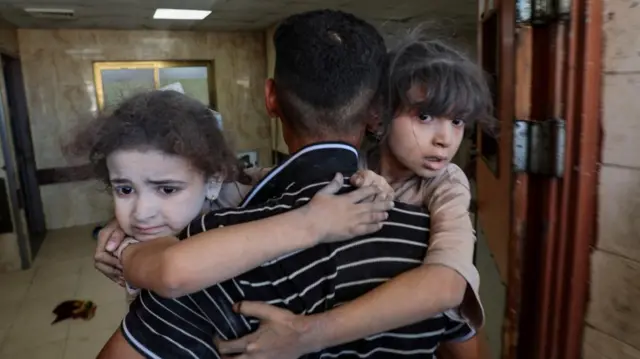 A man carrying two small children whose faces are covered in dust arrive at al-Aqsa Martyrs hospital
