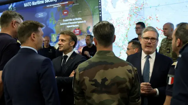 Macron and Starmer, wearing suits, speak witgh military officers and officials.