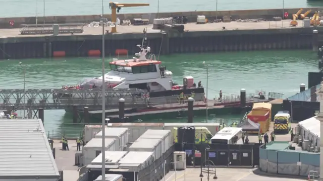 A boat in a dock in greenish water. There's a lot of industrial buildings on the dock, and police mill about