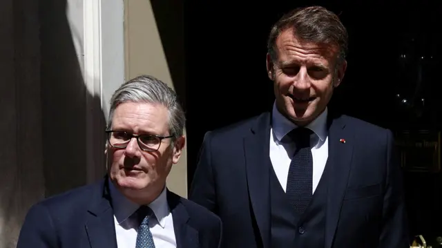UK Prime Minister Keir Starmer (L) in a dark blue suit, white shirt and patterned blue tie steps out of Number 10 Downing Street. Behind him, to his left, is French President Emmanuel Macron in a black suit, black vest, white shirt and black tie, smiling with his eyes partially closed due to the sun shining from in front of him