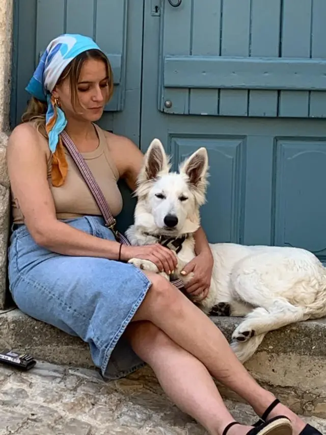 Clemence sits on a step in front of a door with her white god. She’s wearing a blue bandana around her brown hair as well as a brown tank top and a denim skirt. She’s looking at her dog.