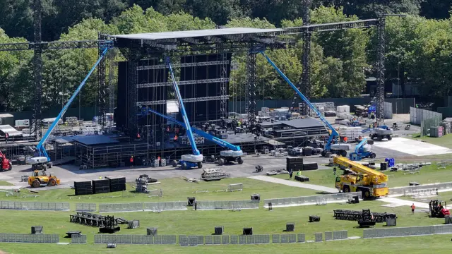 Cranes and other heavy machinery is used to set up a large stage in a wide, open park.