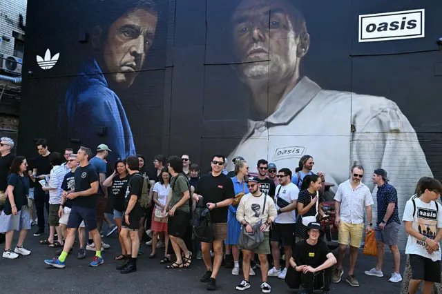 Fans of Oasis, wearing band-branded merchandise, stand in front of a mural depicting Noel and Liam Gallagher and Oasis and Adidas signs