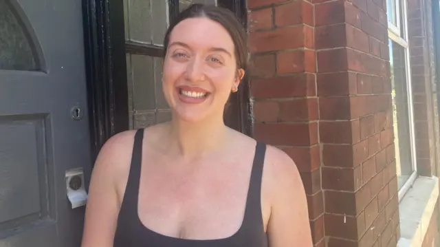 Emily has dark hair pulled back from her face and wears a black vest top