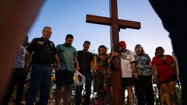 A group of people join hands and bow their heads in prayer in front of a wall of flowers and a large wooden cross