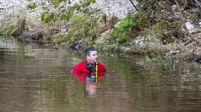 Jack Goodroe Tomball, member of a search and rescue team, looks for missing people near Camp Mystic. He is in the river which is up to his chest and he is wearing a red suit and has swimming goggles on his neck.