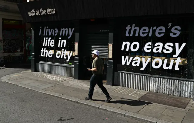 A man in a bucket hat walks past a bar, with the windows adorned with the words "I live my life in the city" and "there's no easy way out"