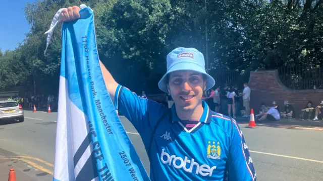 Jorge Arredondo wears a blue Oasis bucket hat, a blue Manchester City football shirt and holds a blue Oasis flag