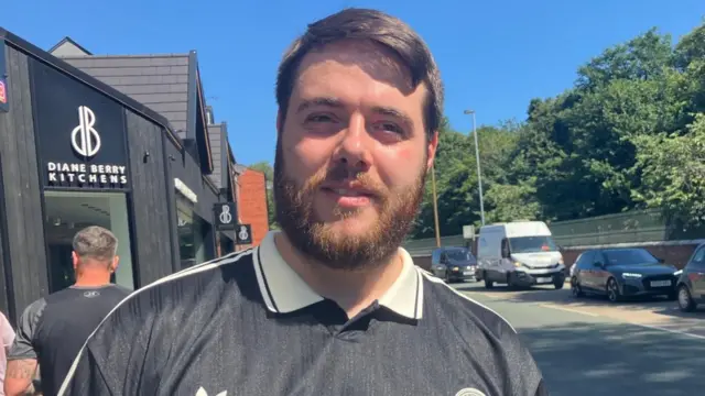 Dylan Matthews is wearing a black Oasis T-shirt and is smiling at the camera.