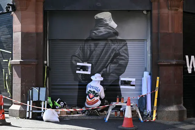An artist paints an image of a man in a bucket hat and a parka coat on the shutters of a shop
