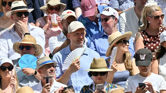 A crowd of spectators watching a tennis match, many wearing caps and hats as well as glasses. Some hold fans.