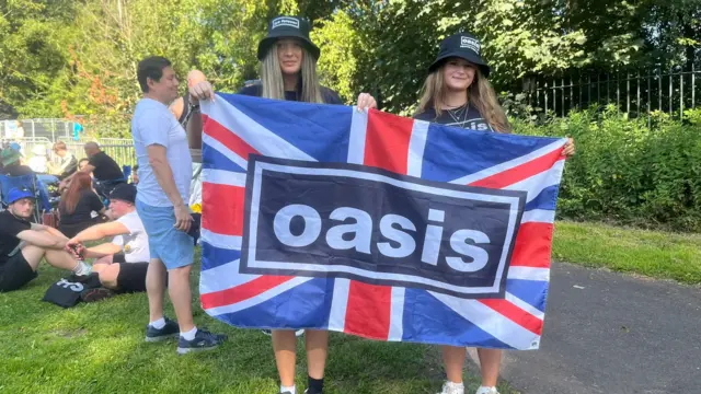 The friends are wearing black bucket hats and are holding a Union Jack Oasis flag
