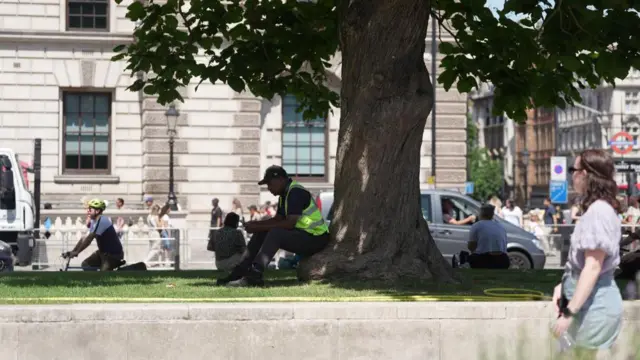 A person sits in the shade at the bottom of a tree in busy and sunny scene in Westminster, London.