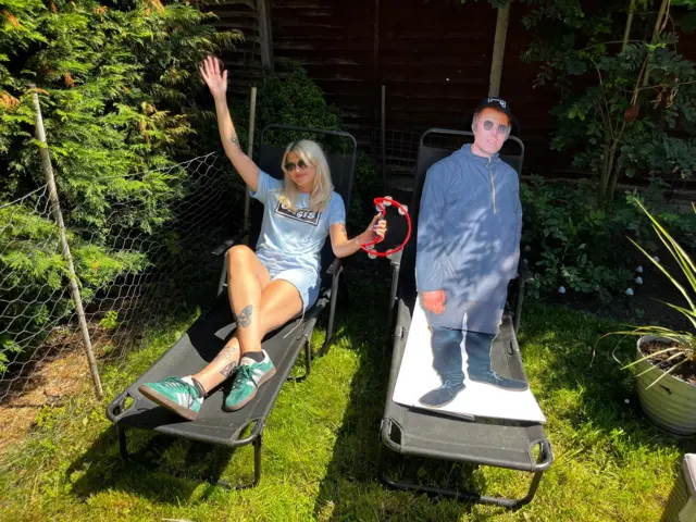 Liv lies on a sun lounger in her back garden. She has shoulder-length blonde hair she wears a blue Oasis T-shirt and matching blue shorts and holds a red halfmoon tambourine. A cardboard cut-out of Liam has been placed on a sun lounger next to her. .
