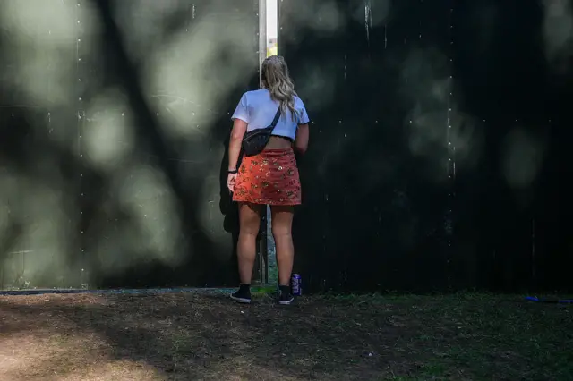 Woman in blue T-shirt and red skirt looks through a gap in the security fencing at the Oasis show in Heaton Park