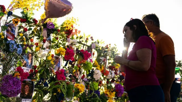 A man and a woman stand by a wall of flowers with pictures of people killed in the flood
