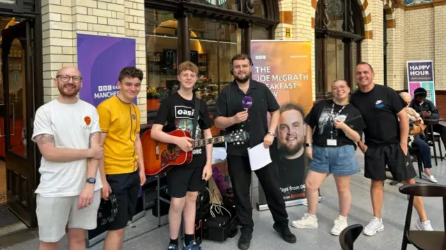 Radio Manchester Breakfast Show host Joe McGrath is speaking to fans at Victoria railway station. He is holding a microphone and is wearing headphones. Two of his colleagues are standing to his right and there three Oasis fans on his left.