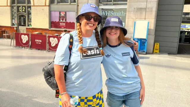 Gayle McPherson and Isla are wearing light blue Oasis T-shirts and bucket hats. They are smiling is this posed photograph in Victoria railway station.