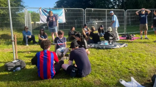 Fans are sitting on the grass outside Heaton Park. There is an Oasis flag hung on the railings and a few of fans are wearing bucket hats
