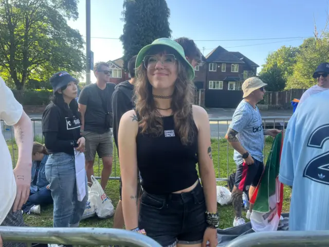 Carley Williams is wearing a green bucket hat and she is smiling. A number of fellow fans can also been seen in the background as they await the opening of Heaton Park.