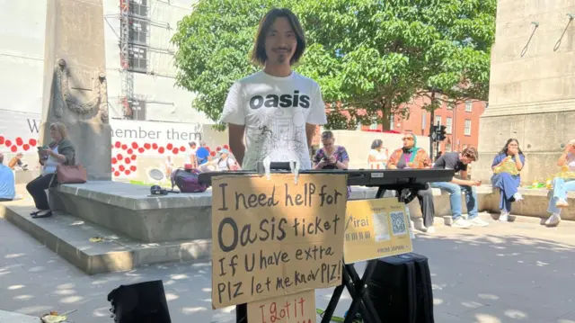 Miraj Ito is standing in front of a sign that reads: "I need help for Oasis ticket."