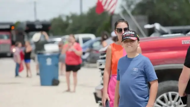A boy wearing a Trump hat smiles in the foreground while other supporters gather in the background of a Texas parking lot
